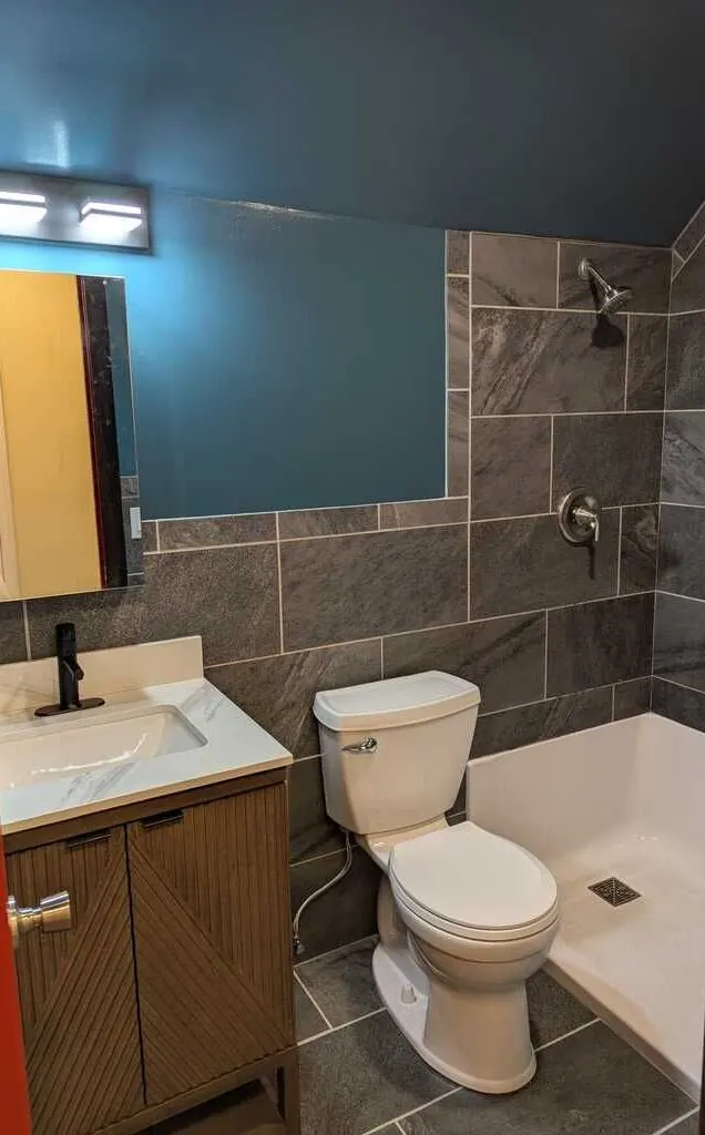 Renovated bathroom with large slate-look wall tile, a shower base, a fluted wood vanity and a black faucet