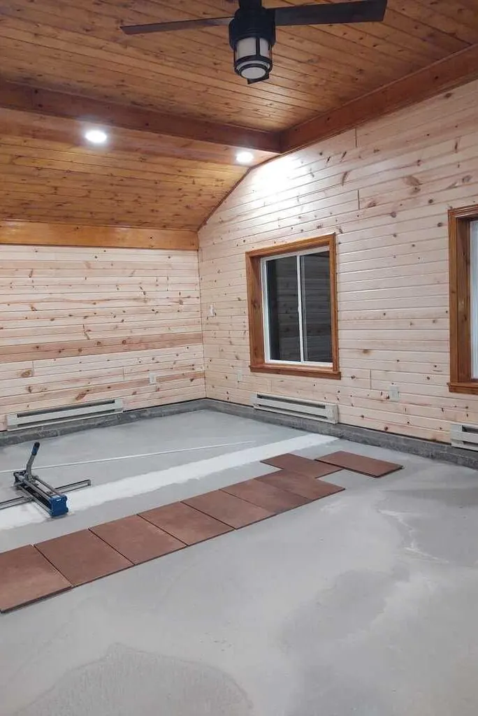 Room mid-build with horizontal pine board walls, a pine ceiling, stained window casing and floor tile being laid
